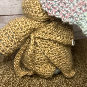 May include: Handmade crocheted doll in a tan outfit with a hood, and a light blue and pink hat. The doll's outfit is tied with a crocheted string. The background is a brown surface.