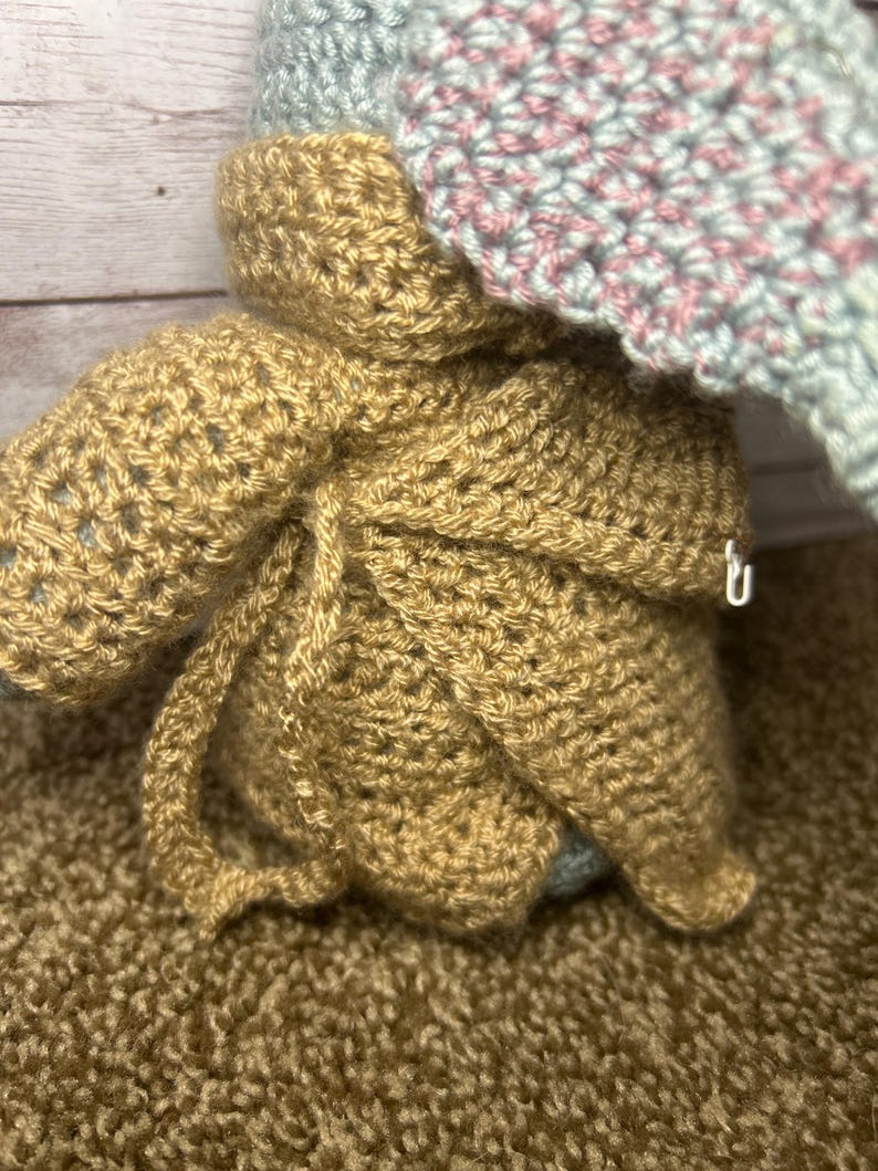 May include: Handmade crocheted doll in a tan outfit with a hood, and a light blue and pink hat. The doll's outfit is tied with a crocheted string. The background is a brown surface.