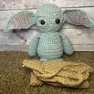 May include: A crocheted figure of a character with large ears, brown eyes, and a tan blanket. The figure is light blue with pink accents inside the ears. The blanket is tan and folded.
