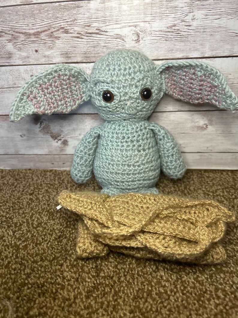 May include: A crocheted figure of a character with large ears, brown eyes, and a tan blanket. The figure is light blue with pink accents inside the ears. The blanket is tan and folded.