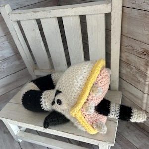 May include: A handmade crochet raccoon toy in black, white, pink, and yellow, resting on a small white wooden chair. The raccoon has a striped tail and a yellow stripe on its head.