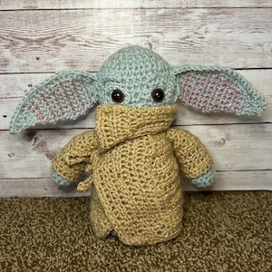 May include: A handmade crochet doll with a light green head and large ears with pink detailing. The doll is wearing a tan robe and has large black eyes. The doll is on a brown carpet with a wooden backdrop.
