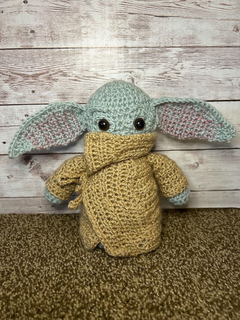 May include: A handmade crochet doll with a light green head and large ears with pink detailing. The doll is wearing a tan robe and has large black eyes. The doll is on a brown carpet with a wooden backdrop.