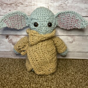 May include: A handmade crochet doll of a character with large ears, brown eyes, and a tan robe. The doll's head and ears are light blue with pink accents. The robe is tan, and the doll is sitting on a brown surface.