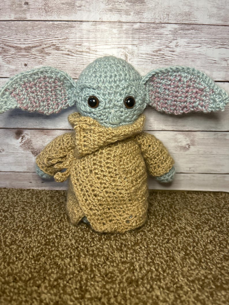 May include: A handmade crochet doll of a character with large ears, brown eyes, and a tan robe. The doll's head and ears are light blue with pink accents. The robe is tan, and the doll is sitting on a brown surface.
