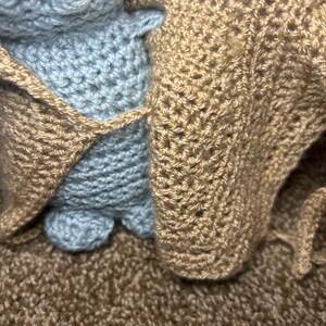 May include: A handmade crochet doll in pale blue, wrapped in a tan-coloured crocheted blanket. The doll's features are simple, with visible stitches and a soft texture. The blanket has a textured appearance.