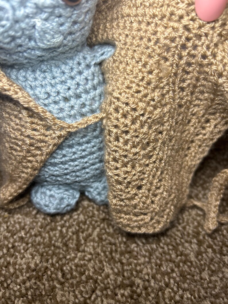 May include: A handmade crochet doll in pale blue, wrapped in a tan-coloured crocheted blanket. The doll's features are simple, with visible stitches and a soft texture. The blanket has a textured appearance.