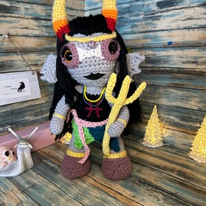 May include: A handmade crochet doll with gray, black, and colorful accents. The doll has large eyes, horns, and a trident. It is wearing a black top with a red symbol and a necklace. The doll is standing on a wooden surface.