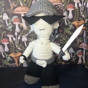 Puede incluir: Muñeca de ganchillo hecha a mano con gorra gris, gafas de sol negras y pantalones negros. La muñeca sostiene una espada blanca. El cuerpo de la muñeca es blanco, con detalles grises y negros. Fondo de pantalla con motivos de setas.