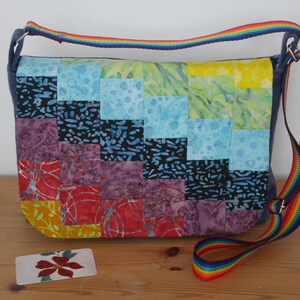 May include: A patchwork crossbody bag with a rainbow strap. The bag is made of square fabric patches in red, purple, blue, and yellow. A small card with a floral design is on the wooden surface. The bag has a rainbow strap.