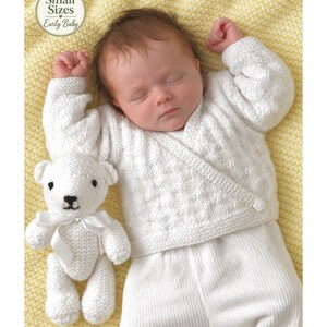 May include: A sleeping infant wearing a white long-sleeved wrap cardigan with a textured knit pattern and a single button closure, paired with white ribbed pants. Beside the infant is a small white knitted teddy bear with a white satin ribbon.