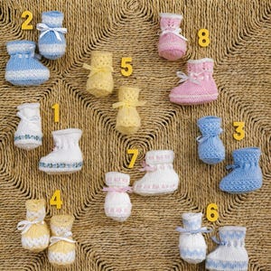 May include: A collection of eight pairs of knitted baby booties in various colors including blue, white, yellow, and pink. Each pair features a ribbon tie at the ankle and is labeled with a yellow number from one to eight, showcasing different textures.