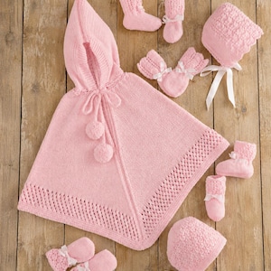 May include: A light pink knitted baby set consisting of a hooded cape with pom-poms, two pairs of booties with white ribbon bows, two pairs of mittens with white ribbon bows, and two bonnets with white ribbon ties, all featuring textured patterns.