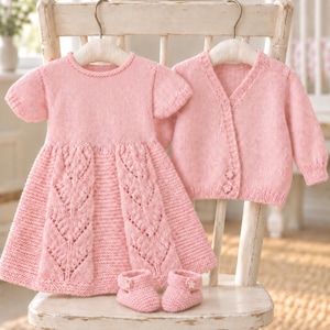 May include: A light pink knitted baby set consisting of a short-sleeved dress with a textured leaf pattern on the skirt, a long-sleeved button-up cardigan, and a pair of matching booties with small flower accents, all displayed on hangers.