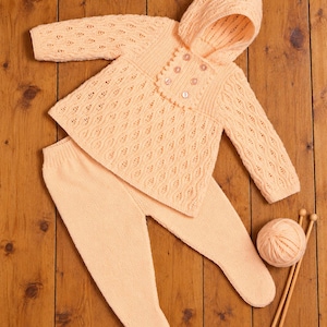 May include: A peach-colored knitted baby outfit consisting of a long-sleeved hooded jacket with a textured lace pattern and four buttons, paired with matching footed leggings. A ball of matching yarn and two knitting needles are included.