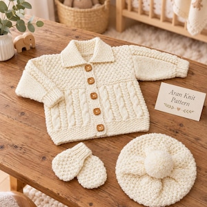 May include: A cream-colored knitted cardigan with a collar, five square wooden buttons, and cable knit detailing. The set includes a pair of matching mittens and a round beret with a central pom-pom. A small card displays the text Aran Knit Pattern.