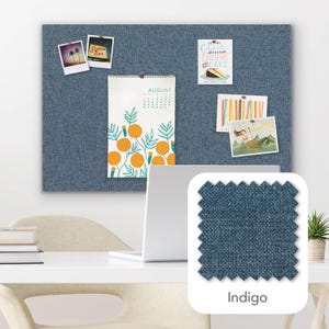 Pu&ograve; includere: Una bacheca in tessuto blu con vari elementi appuntati, tra cui un calendario, foto e una carta. Un campione di tessuto indaco &egrave; mostrato nell'angolo. Un laptop e una piccola pianta sono sulla scrivania bianca.