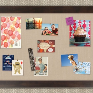 May include: A corkboard with a brown frame and a beige fabric surface. The corkboard is decorated with various photos, cards, and a sticky note that says "Let's make this!" The photos include a watercolor painting of red balloons, a photo of two people in the water, a photo of two people holding a watermelon, a photo of a cupcake, a photo of a woman in a kitchen, and a photo of a Ferris wheel. The cards include a baby shower invitation, a birthday card, and a card with a vintage design.