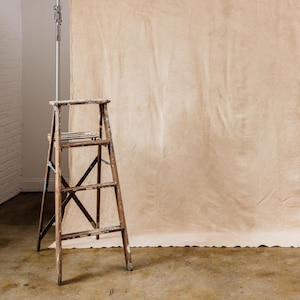 May include: A light beige backdrop hung on a white wall with a wooden step ladder in front of it.