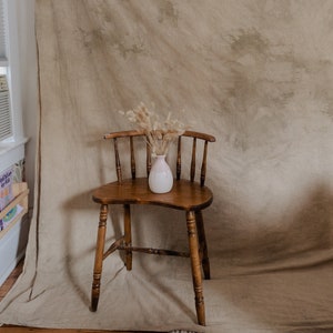 May include: A wooden chair with a curved back and a vase of dried flowers on the seat. The chair is in front of a beige backdrop with brown and white splotches.