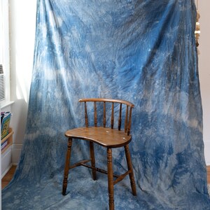 May include: A wooden chair with a curved backrest and four legs sits in front of a large piece of fabric featuring a blue and white tie-dye pattern. The fabric hangs vertically, displaying varied shades of indigo and white in a cloud-like design.
