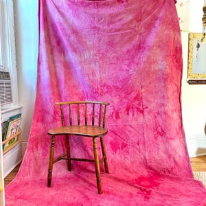 May include: A wooden chair with a spindle back sits in front of a large, vibrant pink backdrop. The backdrop has a tie-dye effect with varying shades of pink and red. The chair is made of wood and has a natural finish.