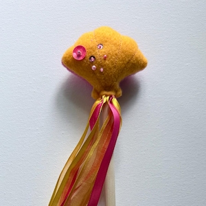 May include: A whimsical yellow felt wand with a cloud-like top decorated with pink sequins and a button. The wand features pink, gold, and orange ribbons and a natural wood handle, ideal for imaginative play.
