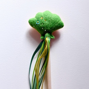 Peut inclure: Une baguette fantaisiste en feutre vert avec un sommet en forme d'arbre orné d'un bouton et de petites perles dorées. Des rubans verts et dorés tombent en cascade de la base, attachés à une cheville en bois naturel.