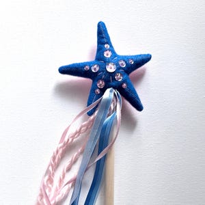 May include: A blue starfish wand with clear and pink rhinestones. The wand has a wooden handle and is adorned with pink, light blue, and dark blue ribbons. A whimsical accessory for imaginative play or themed events.