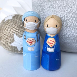 Könnte beinhalten: Zwei Holz-Steckpuppen, die als medizinisches Personal verkleidet sind. Beide Puppen tragen blaue Kittel, weiße Gesichtsmasken und ein rotes Superman-Logo. Die Puppe links trägt eine blaue OP-Haube und hat ein Stethoskop um den Hals. Beide Puppen haben "NHS" auf ihren Kitteln.