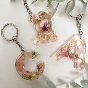 May include: Three resin keychains with floral and gold leaf inclusions. One is a teddy bear shape, and the others are the letters "C" and "A". Each keychain has a silver-colored chain and keyring. The resin is clear with pink and gold accents.