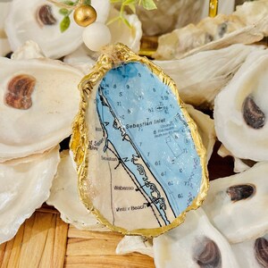 May include: A blue and white nautical map of Sebastian Inlet, Florida, is mounted in a gold-painted oyster shell. The map shows the coastline, waterways, and surrounding areas.