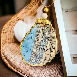 May include: A gold-leafed oyster shell ornament with a map of Naples, Florida inside. The ornament is hanging from a gold and white beaded string.