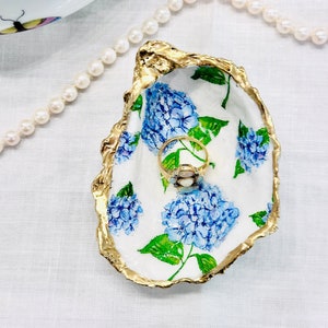 May include: A white ceramic oyster shell ring dish with a gold rim. The dish is decorated with blue and green hydrangeas. A gold ring with a pearl is sitting in the dish.
