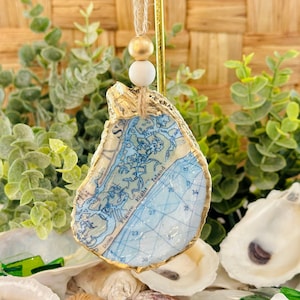 May include: An oyster shell ornament featuring a map design in shades of blue and white. The shell is trimmed with gold paint and has a jute string for hanging, accented with wooden beads. The background includes greenery and other oyster shells.