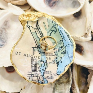 May include: A gold ring with opal stones sits on a decorative oyster shell. The shell is painted with a map of St. Augustine, Florida, and has a gold-painted edge. The ring dish is surrounded by other oyster shells.