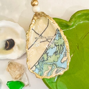 May include: A gold-trimmed oyster shell ornament with a map of Murrells Inlet, South Carolina, inside. The ornament is hanging from a twine string with gold and white beads.