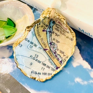 May include: A gold-leafed oyster shell ornament with a map of Indian Rocks Beach, Florida, printed on the inside. The map includes details like the location of a lighthouse, a beach, and a channel.