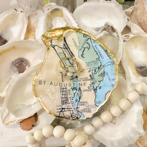 May include: An oyster shell ring dish featuring a map of St. Augustine, Florida, and a gold ring with white gemstones. The dish has a gold edge and is surrounded by other oyster shells and a beaded garland.