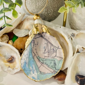 May include: A gold-painted oyster shell ornament with a map of St. Simons Island printed inside. The ornament is surrounded by other oyster shells and seashells.