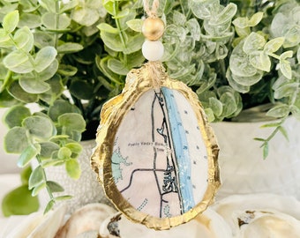 May include: Gold-painted oyster shell ornament with a map of Ponte Vedra Beach, Florida inside. The ornament has a white bead and gold bead hanging from a twine string.