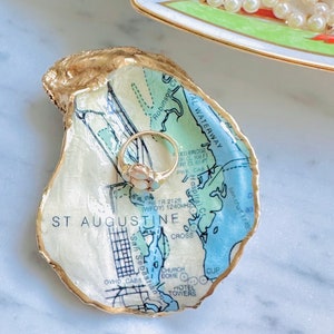May include: An oyster shell trinket dish with a map of St. Augustine, Florida, featuring gold edging. A gold ring with a white stone sits inside the dish. A decorative dish with a pearl necklace is in the background.