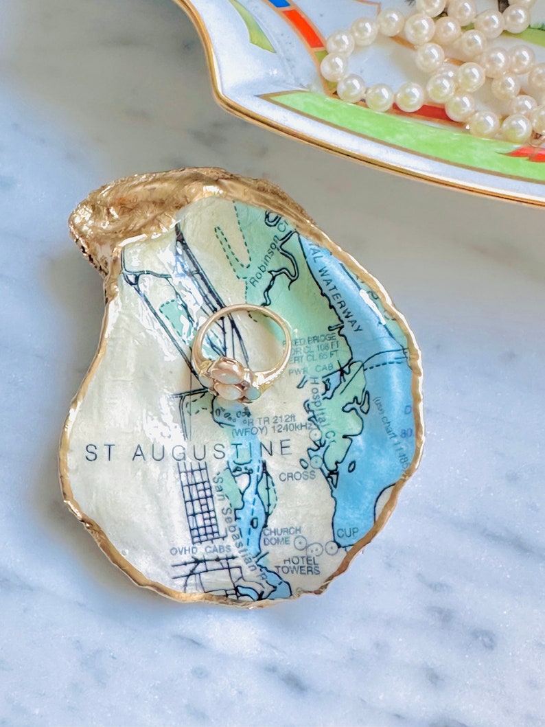 May include: An oyster shell trinket dish with a map of St. Augustine, Florida, featuring gold edging. A gold ring with a white stone sits inside the dish. A decorative dish with a pearl necklace is in the background.