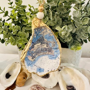 May include: Gold oyster shell ornament with a map of Marco Island, Florida inside. The ornament is hanging from a string with a white and gold bead. The ornament is surrounded by other oyster shells and white beads.