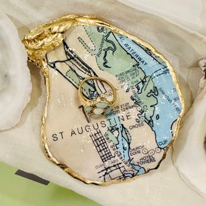 May include: An oyster shell ring dish with a gold-painted rim. The interior features a map of St. Augustine, Florida, with a gold ring with a cluster of stones. The dish is on a white surface.