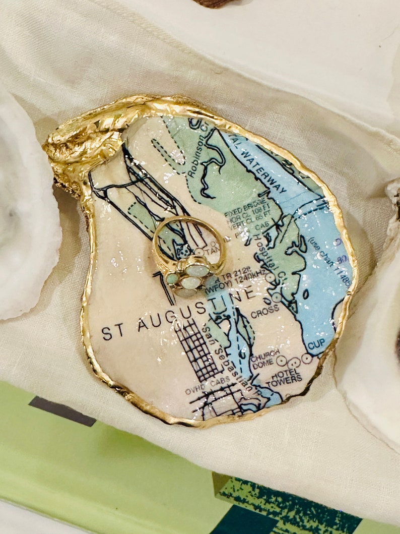 May include: An oyster shell ring dish with a gold-painted rim. The interior features a map of St. Augustine, Florida, with a gold ring with a cluster of stones. The dish is on a white surface.