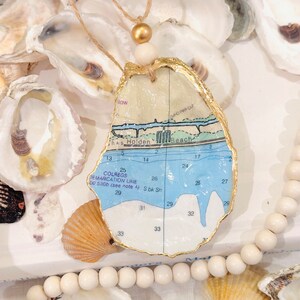 May include: A gold-leafed oyster shell with a map of a coastal area inside. The map shows the words "Holden Beach" and "Colregs Demarcation Line 100 5300 (see note A) Sbk Sh". The oyster shell is hanging from a white and gold cord with a wooden bead.