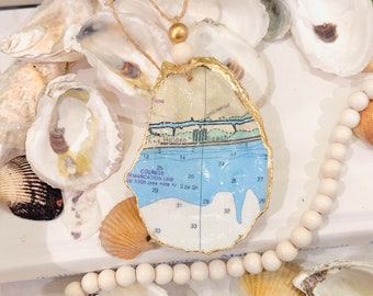 May include: A gold-leafed oyster shell with a map of a coastal area inside. The map shows the words "Holden Beach" and "Colregs Demarcation Line 100 5300 (see note A) Sbk Sh". The oyster shell is hanging from a white and gold cord with a wooden bead.