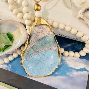 May include: A gold-painted oyster shell with a nautical map inside. The shell is hanging from a white beaded garland.