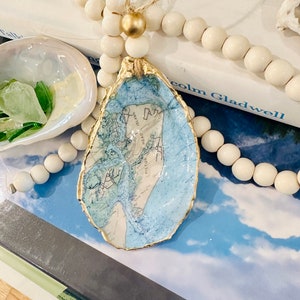 May include: A gold-painted oyster shell ornament with a blue and white map inside. The ornament is hanging from a string of white wooden beads.